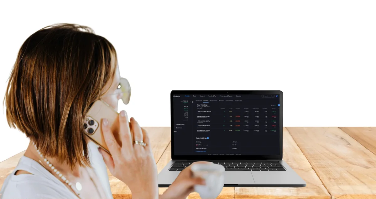 A woman holds her phone to her ear and talks to marlo while looking at her brokerage holdings on her laptop.